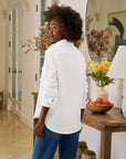 A woman with curly hair stands indoors, looking over her shoulder. She wears the Frank & Eileen Eileen Sweatshirt Button Up with blue jeans. A table with flowers, peaches, and a lamp is in the background.