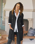 A woman with curly hair smiles indoors, wearing the Frank & Eileen Sweatshirt Trench Coat in black over a white shirt as she leans on a wooden table near a fireplace with a striped pillow.
