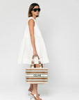 A woman wears the SUNCOO Conely Dress, a relaxed fit, sleeveless white summer dress, paired with white heeled sandals and large black sunglasses. She holds a striped Celine tote bag and stands against a plain white background.