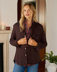 A woman with long blonde hair stands indoors wearing the Frank & Eileen Eileen Button Up Shirt F25 under a dark brown jacket with blue jeans. She looks at the camera, hands on her jacket; a plant and candle enhance the cozy California vibe.
