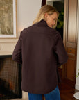 A woman with blonde hair, seen from behind, wears the Frank & Eileen Eileen Button Up Shirt F25 in a relaxed fit along with blue jeans, standing indoors by a fireplace and wooden cabinets.