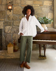 A person with curly hair, wearing a relaxed-fit, 100% cotton Oona Continuous Button-Up Shirt by Frank & Eileen and green pants, stands with one hand resting on a piano in a room with stone walls. There is a plant on the piano and two large vases on the floor nearby. A lit wall sconce adds warm light to the scene.