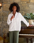 A person stands indoors, leaning against a piano in front of a stone wall. They wear the Oona Continuous Button-Up Shirt from Frank & Eileen, which is a relaxed fit, 100% cotton white shirt, along with green pants. Their curly hair is loose, and they smile slightly. The top of the piano holds a vase with green foliage and a few small decorative items.