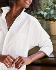 A person with curly hair is leaning against a wooden surface. They are wearing the Oona Continuous Button-Up Shirt made from 100% cotton in white, by Frank & Eileen, with the collar popped up and sleeves rolled. The person also wears gold rings and a delicate gold necklace. A potted plant is blurred in the background.