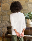 A person with curly hair is standing with their back turned, facing a stone wall and an upright piano adorned with decorative plants on top. They are wearing a Frank & Eileen Oona Continuous Button-Up Shirt made from 100% cotton and green pants, with their hands clasped behind their back.