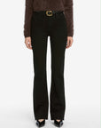 A person wears black bootcut jeans, a fuzzy dark sweater, and pointed black heels, styled with FRAME's The Organic Buckle Belt featuring a gold buckle, all set against a white background.