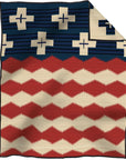 The Jacquard Robe by Pendleton/Babbitt's Wholesale is a woven wool blanket featuring navy and cream crosses on dark blue and wavy red and cream stripes, with a folded corner showing the reverse. Inspired by Native American designs, made in USA.