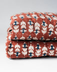 Two folded Blossom Block Print Quilts by Faire, featuring a rust background with a repeating white and blue leaf pattern, stacked on a light surface—perfect for adding bohemian bedding style to your space.