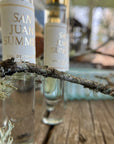 Close-up of two Faire San Juan Roll On perfume bottles on wood, with a lichen-covered twig in front and a blurred backdrop—evoking the cedar and spice scents of the San Juan Islands.