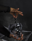 A hand with dark nails drops a black crystal into the Accent Decor Reaper Pot, a skull-shaped glass terrarium shrouded in smoke and cobwebs—ideal for spooky decor on a mysterious dark background.