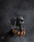 A hand with long, decorated nails holds the Accent Decor Reaper Pot, a transparent skull-shaped terrarium filled with dark liquid against a smoky backdrop—perfect for adding witchy decor vibes.