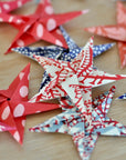 Faire’s Origami Star Garland features red, blue, and white patterned stars made from quality origami paper, artfully displayed on a light wooden surface.