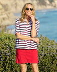 A woman wearing sunglasses and a short-sleeved Patty Popover SS Henley by Frank & Eileen stands smiling outdoors in front of green bushes with a blurred seaside in the background.