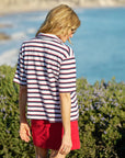 A woman with blonde hair stands outdoors near greenery and the ocean, wearing a Frank & Eileen Patty Popover SS Henley and a red skirt. The coastal background features blue water and lush scenery.