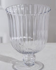 The Oster Vase M by Accent Decor, a clear glass vase with ribbed detailing, a wide flared rim, and a short pedestal base, is displayed on a white surface against a light background.