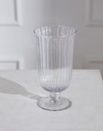 The Oster Vase S by Accent Decor—a clear, fluted glass vase—stands on a white surface, casting a faint shadow. Its elegant, handmade-inspired lines are highlighted against softly lit, white paneled walls in the background.