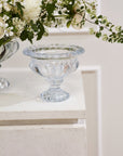 The Hathaway Compote S by Accent Decor, a clear vintage glass piece with a wide rim and decorative base, sits on a white textured surface beside a floral arrangement of white flowers and green leaves.