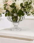 The Hathaway Compote M by Accent Decor holds a bouquet of white and pale peach flowers with green leaves, displayed on a white surface against a light background.