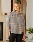 A blonde woman wears the Frank & Eileen Eileen Button Up Shirt F25 with bust-flattering buttons and black pants, standing indoors by a fireplace, near a table with a plant and magazines.
