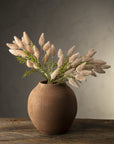 The Indaba Pussytail Grass Spray, featuring fluffy beige dried stems and green faux plants, is arranged in a round textured brown vase on a rustic wooden table set against a softly lit neutral background.