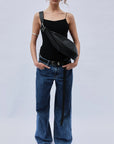 A person wearing sunglasses, a black top, wide-leg blue jeans, and black pointed boots stands against a plain background. With hands in pockets, they sport the Myra DW Belt by Janessa Leone. Their large crossbody bag is made of black regenerative leather with artisan crafted silver hardware.