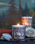 A lit G&W Soy Candle by Faire from the Happy Place Collection sits on a red book beside stones, dried botanicals, and another tin. A painted landscape with trees and mountains creates a sense of nostalgic joy.