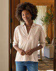 A woman with curly hair wears the Frank & Eileen Patrick Popover Henley, a beige and white striped cropped long-sleeve shirt, paired with blue jeans. She stands indoors, smiling gently, with a blurred living room in the background.
