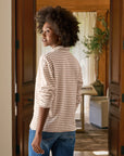 A woman with curly hair stands indoors, sunlight streaming behind her, wearing the Patrick Popover Henley by Frank & Eileen—a striped, cropped long-sleeve shirt—paired with jeans and looking over her shoulder.
