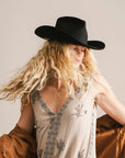 A woman with long, wavy blonde hair wears the Faire Charlie Western Hat and a light embroidered sleeveless dress, with a brown jacket draped over her arms, standing against a plain background.