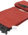 The Rust Red Baja Thunderbird Mexican Blanket by Faire is a handmade, woven yoga blanket from Mexico, featuring black and beige patterns, black fringed edges, and an attached tag for authentic style and warmth.