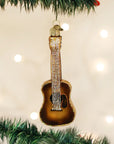 The Old World Ornaments guitar-shaped ornament by Faire, featuring brown and gold details with sparkling strings, hangs on a green Christmas tree branch among other decorations against a backdrop of warm white lights.
