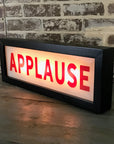 The Faire Lighted Sign is a rectangular lightbox displaying "APPLAUSE" in bold red letters, illuminated against a rustic brick wall background and set on a wooden surface.