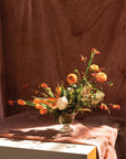 The Camilla Compote by Accent Decor, a glass vase with a 5.5-inch opening, displays orange tulips, yellow and orange flowers, and greenery on a pink-draped table against a soft brown backdrop in warm sunlight.