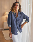 A woman with curly hair, wearing the Eileen 26 relaxed fit shirt by Frank & Eileen and white pants, stands smiling by a window with sheer curtains in a cozy, softly lit room.