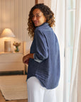 A woman with curly hair, wearing the Frank & Eileen Eileen 26 relaxed fit blue denim shirt and white pants, stands indoors by a window, smiling over her shoulder in a softly lit, neutral-toned room with a lamp on the dresser.