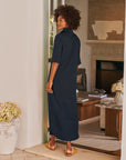 A woman with curly hair stands barefoot in a doorway, wearing the Frank & Eileen Emma Popover Henley Dress in navy. She looks over her shoulder, revealing a cozy living room behind her.