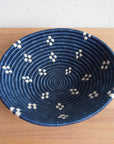 The Kabaya Large Bowl by Amsha - Faire is a round blue sisal woven basket with a coiled design and small white geometric patterns on both sides, shown on a light wooden surface.
