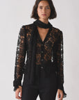 A woman wears The Bow Tie Shirt by Cissa—a sheer black lace blouse with long sleeves and a scalloped hem—paired with black pants and a matching lace scarf, standing against a plain white background.