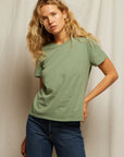 A woman with long wavy blonde hair wears the Perfectwhitetee Harley SS Boxy Crew in light green and mid-rise denim. She stands before a plain beige fabric backdrop, one hand in her pocket, looking at the camera.