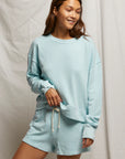 A person with long hair smiles while wearing the Layla Terry Short from Perfectwhitetee, paired with a light blue sweatshirt. Crafted from soft French terry cotton, they stand confidently before a light fabric backdrop, hand resting on their sweatshirt hem, showcasing effortless comfort.