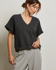 A woman with brown hair in a low bun wears the Perfectwhitetee French Terry SS Crew and light gray drawstring pants, standing against a plain light wall, looking slightly down and to the side.