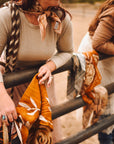 A woman in a tan long-sleeve shirt and Faire's Rustic Wild Rose Western Silk Scarf leans on a metal fence, holding warm-toned patterned fabrics—the perfect cowgirl accessory. Another person stands beside her outside, likely at a ranch or farm.
