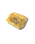The Faire Fashion Hair Clip is hand-painted yellow to resemble a stick of butter, featuring black text and lines reading "Grade AA Butter" and "Net Wt. 4 oz (113g)." The clip casts a shadow on a white background.