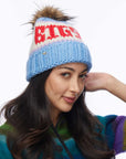 A woman with long dark hair wears the Faire Knit Ski Hat, a luxury merino wool beanie with a faux fur pom pom, paired with a striped multicolored sweater, posing against a plain background.