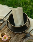 An Indaba 4-7122 Stripe Resist Napkin is neatly folded in a dark bowl on a matching plate, with fork, spoon, and knife set on a woven placemat atop an outdoor table against a grassy backdrop.
