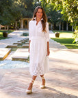 A woman stands outdoors near a pool, wearing the Frank & Eileen Gracie Midi Eyelet Dress with ankle-tie espadrilles. Green trees and a building are in the background.