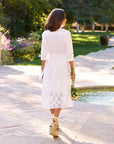 A woman wearing the Frank & Eileen Gracie Midi Eyelet Dress stands by a garden pool, holding a bouquet of flowers behind her back.