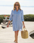 A woman in the Frank & Eileen MARY Shirtdress S25, styled with sunglasses, sandals, and a woven straw bag, strolls a beachside boardwalk framed by flowers and greenery.