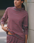 A woman in Donni’s Striped Jersey Turtleneck, featuring red, white, and blue horizontal stripes, stands against a light wall and looks to the side.
