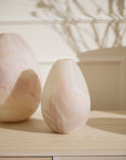A smooth, pale pink Gasparo Vase by Accent Decor with natural marble swirls sits on a light wooden surface, while another handcrafted vase appears in the background amid leafy shadows on the wall.